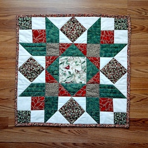 May include: A square quilted table topper with a festive design. The quilt features a central square with two red birds on a white background, surrounded by green, red, and white geometric shapes. The border is a dark red with a floral pattern.