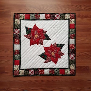 May include: Square quilted table runner featuring two red poinsettia flower appliqués with green leaves on a white background. The border is made of various patterned fabric squares in red, green, and white. The runner is approximately 20 inches square.