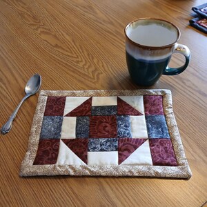 Könnte beinhalten: Ein gequilteter Stoffuntersetzer mit einem geometrischen Muster in Rot-, Weiß- und Blautönen. Der Untersetzer befindet sich auf einem Holztisch mit einem Löffel und einer Tasse.
