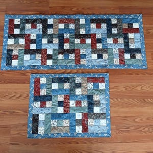 Può includere: Due tovagliette trapuntate con un design patchwork. Le tovagliette rettangolari presentano un mix di tessuti blu, rossi e dai toni neutri disposti a blocchi. Un bordo floreale blu incornicia i bordi.