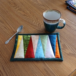 May include: A colorful quilted coaster with a geometric pattern of triangles in shades of green, yellow, red, blue, and white. The coaster has a dark green border and is sitting on a wooden table.