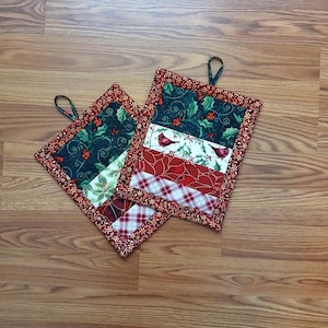 May include: Two square Christmas-themed pot holders with a loop for hanging. The pot holders feature a patchwork design with red, green, and white fabrics. One section has a cardinal bird print, and another has a plaid pattern.