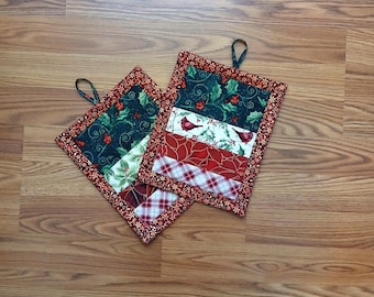 Könnte beinhalten: Zwei quadratische Topflappen im Weihnachtsthema mit einer Schlaufe zum Aufhängen. Die Topflappen haben ein Patchwork-Design mit roten, grünen und weißen Stoffen. Ein Abschnitt hat einen Kardinalvogel-Druck, ein anderer ein Karomuster.