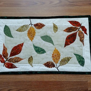 Op de afbeelding: Een rechthoekige stoffen placemat met een crèmekleurig middenpaneel met een subtiel marmerpatroon, versierd met geappliqueerde herfstbladeren in tinten rood, oranje, geel en groen, omlijst door een donkergroene stoffen rand.