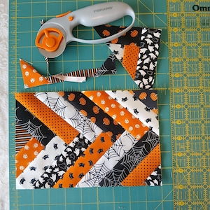 Pode incluir: Um patchwork de tiras de tecido com tema de Halloween em preto, laranja e branco, com padr&otilde;es de ab&oacute;boras, fantasmas e teias de aranha. Um cortador rotativo repousa sobre o tecido, com tiras cortadas por perto. O tecido est&aacute; sobre uma base de corte verde.