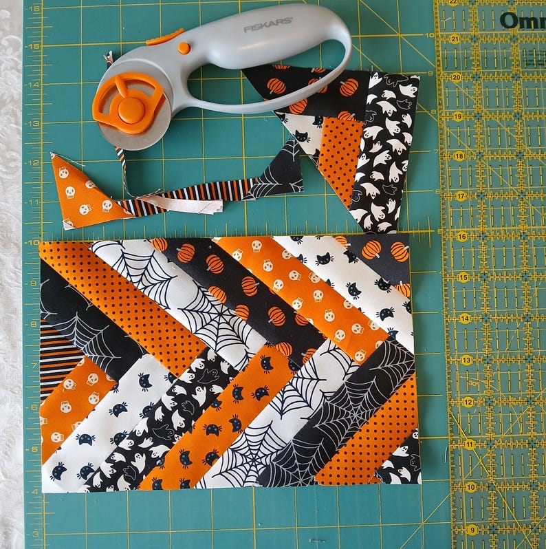 Pode incluir: Um patchwork de tiras de tecido com tema de Halloween em preto, laranja e branco, com padr&otilde;es de ab&oacute;boras, fantasmas e teias de aranha. Um cortador rotativo repousa sobre o tecido, com tiras cortadas por perto. O tecido est&aacute; sobre uma base de corte verde.