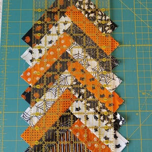 Pode incluir: Um design de patchwork com tiras de tecido com tema de Halloween em laranja, preto e branco, dispostas em zigue-zague. Uma r&eacute;gua transparente com medidas em cent&iacute;metros &eacute; colocada sobre o tecido para cortar e medir. O fundo &eacute; uma base de corte verde.