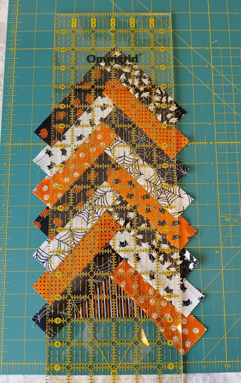 Pode incluir: Um design de patchwork com tiras de tecido com tema de Halloween em laranja, preto e branco, dispostas em zigue-zague. Uma r&eacute;gua transparente com medidas em cent&iacute;metros &eacute; colocada sobre o tecido para cortar e medir. O fundo &eacute; uma base de corte verde.