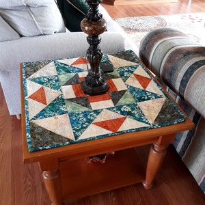 Puede incluir: Una mesa auxiliar de madera con un camino de mesa acolchado en tonos verdes, naranjas y blancos. La mesa tiene un estante inferior y patas torneadas.