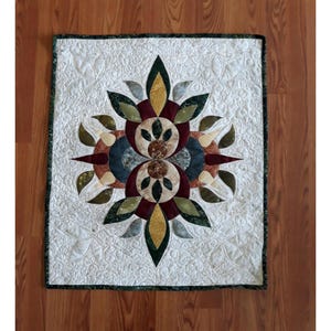 May include: A square, quilted wall hanging with a central floral design. The design features layered fabric in shades of green, burgundy, gold, and blue, set against a white background with leaf-shaped quilting. The border is dark green.