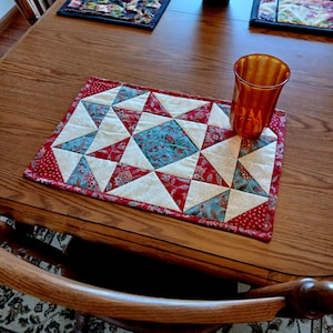 Può includere: Un sottopentola trapuntato fatto a mano con un motivo a stella. Il sottopentola presenta tessuti rossi, bianchi e verde acqua. Un bicchiere color ambra è appoggiato sul sottopentola. Il sottopentola è su un tavolo di legno.