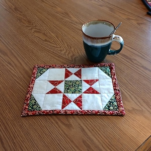 以下が含まれることがあります： 赤、白、緑の星柄の手作りキルトコースター。コースターは長方形で、赤い花の縁取りがあります。銀のスプーンが入ったティールマグがコースターの上に置かれ、木製のテーブルの上に置かれています。