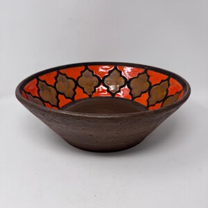 May include: A brown ceramic bowl with a textured exterior. The interior features a vibrant orange band with a repeating pattern of brown quatrefoil shapes. The bowl has a wide, shallow design.