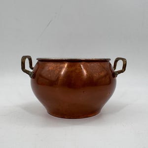 May include: A round copper pot with a smooth, polished finish and a metallic sheen. The vessel features two small, curved brass-toned handles attached to opposite sides and a silver-toned metal rim around the top opening.