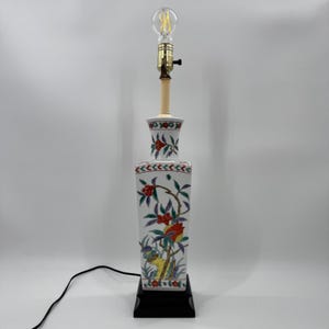 May include: A tall, square-shaped ceramic table lamp featuring a white base decorated with hand-painted floral motifs in red, green, and blue. The lamp has a brass-colored socket, a clear glass light bulb, and a solid black square base with a black cord.