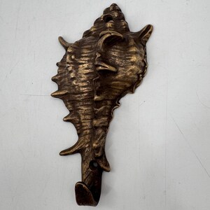 May include: Bronze-colored seashell wall hook. The hook is shaped like a conch shell with intricate details and a curved hook at the bottom. The hook is made of metal and has a textured surface. The design is reminiscent of coastal decor.