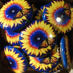 May include: A collection of colorful round buttons featuring a sunflower design with a butterfly in the center. The sunflowers have yellow petals, a blue center, and a white dot in the center. The butterflies are orange and black.