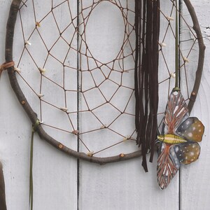 May include: A brown dreamcatcher with a woven net, brown feathers, and a brown leather tassel hanging from a branch. A painted butterfly is attached to the bottom of the dreamcatcher.