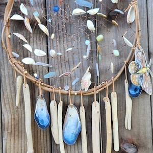 May include: A dreamcatcher with a brown wooden hoop, decorated with seashells, beads, and a blue butterfly. The dreamcatcher has several long, thin, brown wooden strands hanging from the bottom.