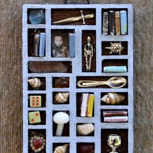 May include: A small, light blue, divided wooden box filled with miniature objects. Items include tiny books, a miniature sword, a photo, a skeleton figurine, seashells, dice, a spoon, and a small chalice. The box is mounted on a wooden surface.