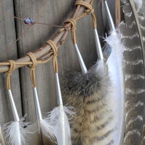 May include: Close-up of a dreamcatcher with brown leather wrappings and feathers. The feathers are white, brown, and black with a striped pattern. The dreamcatcher is hanging on a wooden surface.
