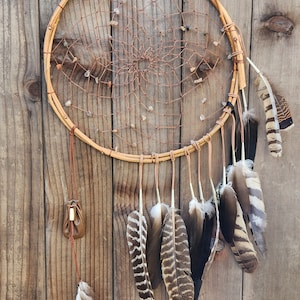 May include: A brown dreamcatcher with a copper wire web and small stones hanging from it. The dreamcatcher is adorned with a variety of brown feathers.