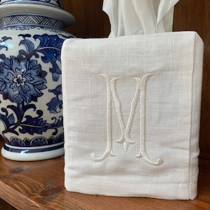 Op de afbeelding: Een vierkante witte stoffen tissueboxhoes met een centraal geborduurde letter M in bijpassende witte draad. De hoes is gemaakt van een getextureerd geweven materiaal en is ontworpen om over een standaard rechthoekige tissuebox te passen.