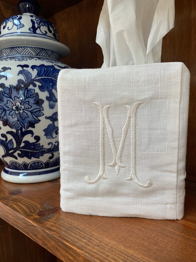 Op de afbeelding: Een vierkante witte stoffen tissueboxhoes met een centraal geborduurde letter M in bijpassende witte draad. De hoes is gemaakt van een getextureerd geweven materiaal en is ontworpen om over een standaard rechthoekige tissuebox te passen.