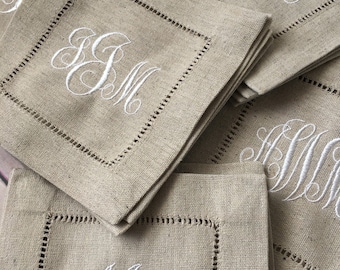 May include: A collection of square beige linen napkins featuring a white embroidered monogram in the centre. Each napkin has a decorative hemstitch border detail surrounding the fabric. The white thread creates a contrast against the natural tan fabric.