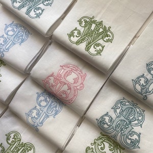 May include: A collection of folded white fabric napkins, each featuring a large, intricate embroidered monogram letter in various colors including light blue, sage green, teal, and soft pink, showcasing detailed scrollwork and ornate flourishes.