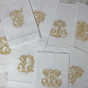 May include: A collection of white rectangular linen napkins, each featuring a distinct gold-colored embroidered monogram letter in an ornate, swirling script style. The napkins have a horizontal hemstitch detail near the bottom edge.