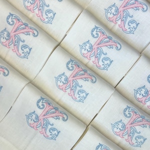 May include: A collection of white fabric napkins arranged in a grid, each featuring an identical embroidered monogram of the letter K. The letter is stitched in a pink thread with intricate blue scrollwork detailing surrounding the character.