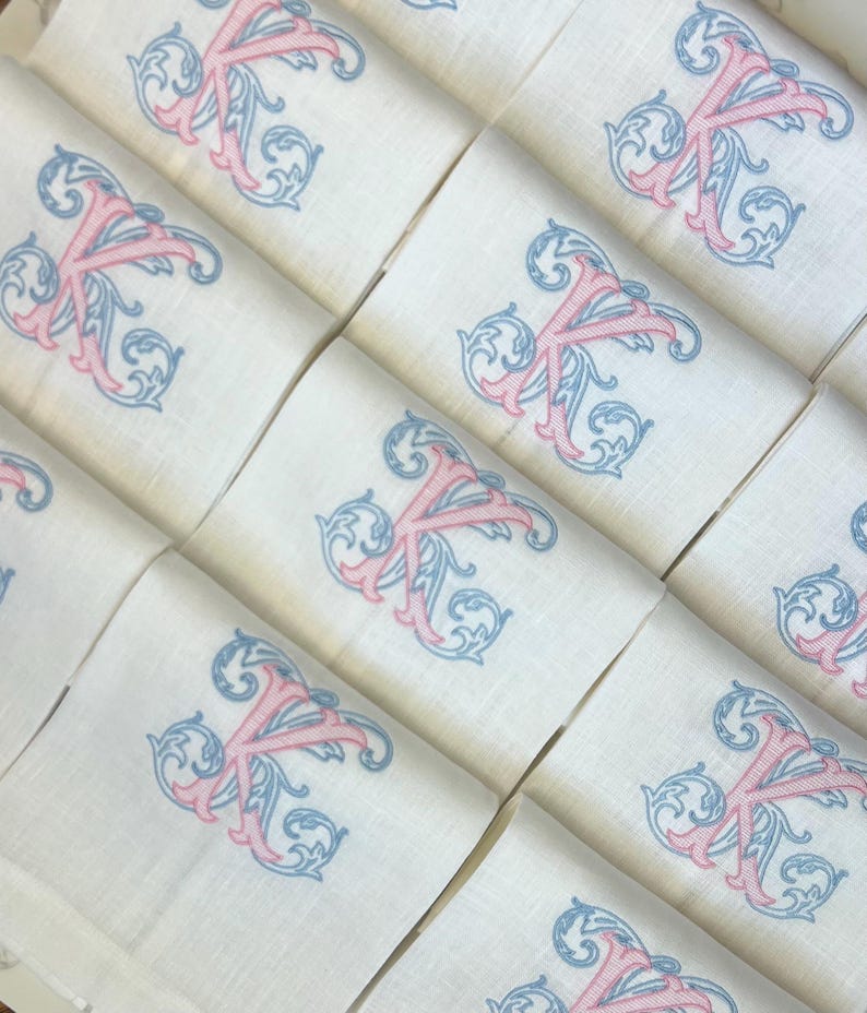 May include: A collection of white fabric napkins arranged in a grid, each featuring an identical embroidered monogram of the letter K. The letter is stitched in a pink thread with intricate blue scrollwork detailing surrounding the character.