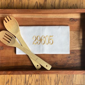 May include: A white kitchen towel with the number 29605 embroidered in gold thread, resting on a wooden serving tray with two wooden spoons.