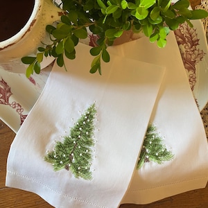 Puede incluir: Dos servilletas de lino blanco con árboles de Navidad bordados en verde decorados con cuentas blancas.