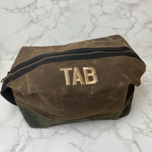 Puede incluir: Una bolsa de lona marrón con cremallera negra y las letras bordadas "TAB" en dorado. La parte inferior de la bolsa es de color verde oscuro. La bolsa está sobre una superficie de mármol.
