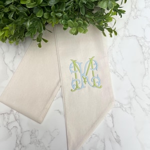 May include: A beige sash with an embroidered monogram in blue and green. The sash is folded and lies on a marble surface, with a green leafy wreath in the background. The monogram is the letter "M".