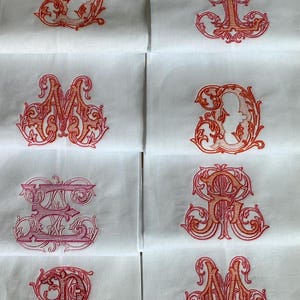 May include: A collection of white fabric squares, each featuring a distinct, ornate embroidered letter in shades of pink and orange. The letters are rendered in a decorative, swirling script style, centered on each individual white textile piece.