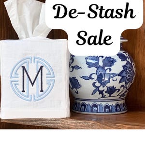 May include: A white linen tissue box cover featuring a light blue and navy blue embroidered monogram letter M inside a circular frame, positioned next to a blue and white floral patterned ceramic ginger jar with the text De-Stash Sale.