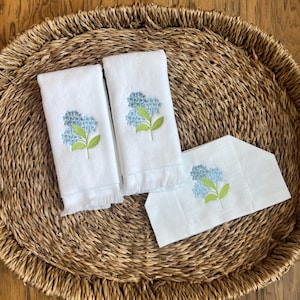 May include: White hand towels and a guest towel with embroidered blue hydrangea flowers and green leaves. The towels have a fringed edge and are arranged on a woven oval tray, creating a spa-like aesthetic.