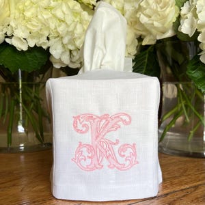 May include: A white linen tissue box cover with a pink embroidered letter "K" in an ornate script. The tissue box is square and has a stack of white tissues emerging from the top. The background features white flowers.