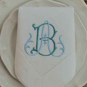 Può includere: Un tovagliolo di stoffa bianco con un monogramma ricamato delle lettere A e B in tonalità verde acqua e azzurro. Il ricamo presenta intricati ghirigori e un dettaglio a stella centrale, con un bordo decorativo a giorno lungo il margine.