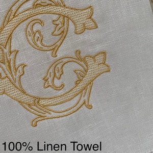 May include: A white 100% linen towel featuring an ornate, gold-colored embroidered letter C with intricate scrollwork and leaf-like details. The fabric has a visible woven texture and a decorative hemstitch border along the bottom edge.