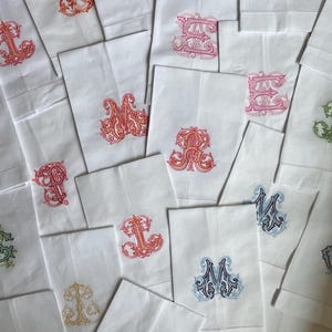 May include: A collection of white rectangular fabric napkins, each featuring a unique, ornate embroidered monogram in various colors including red, orange, pink, blue, green, and gold. The embroidery displays intricate, swirling letter designs.