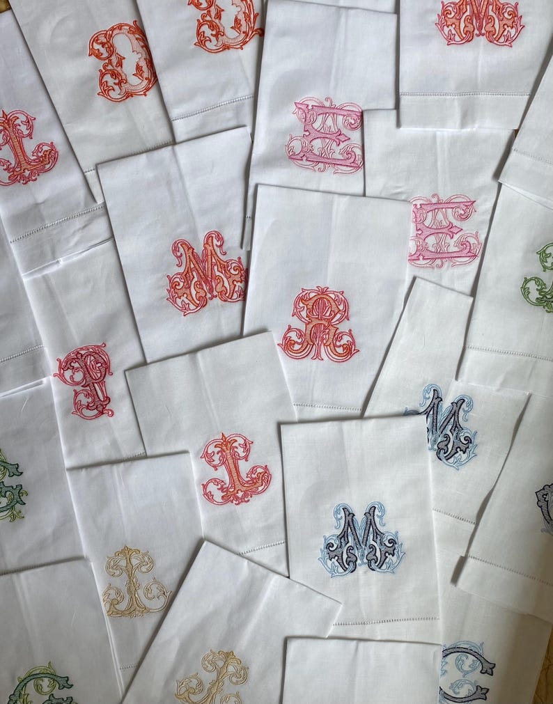 May include: A collection of white rectangular fabric napkins, each featuring a unique, ornate embroidered monogram in various colors including red, orange, pink, blue, green, and gold. The embroidery displays intricate, swirling letter designs.