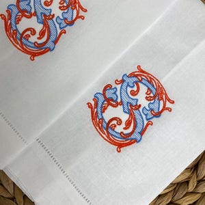 May include: Two white linen napkins featuring an embroidered monogram letter S in a decorative script style. The embroidery uses a combination of light blue thread for the letter and vibrant orange thread for the surrounding ornate scrollwork.