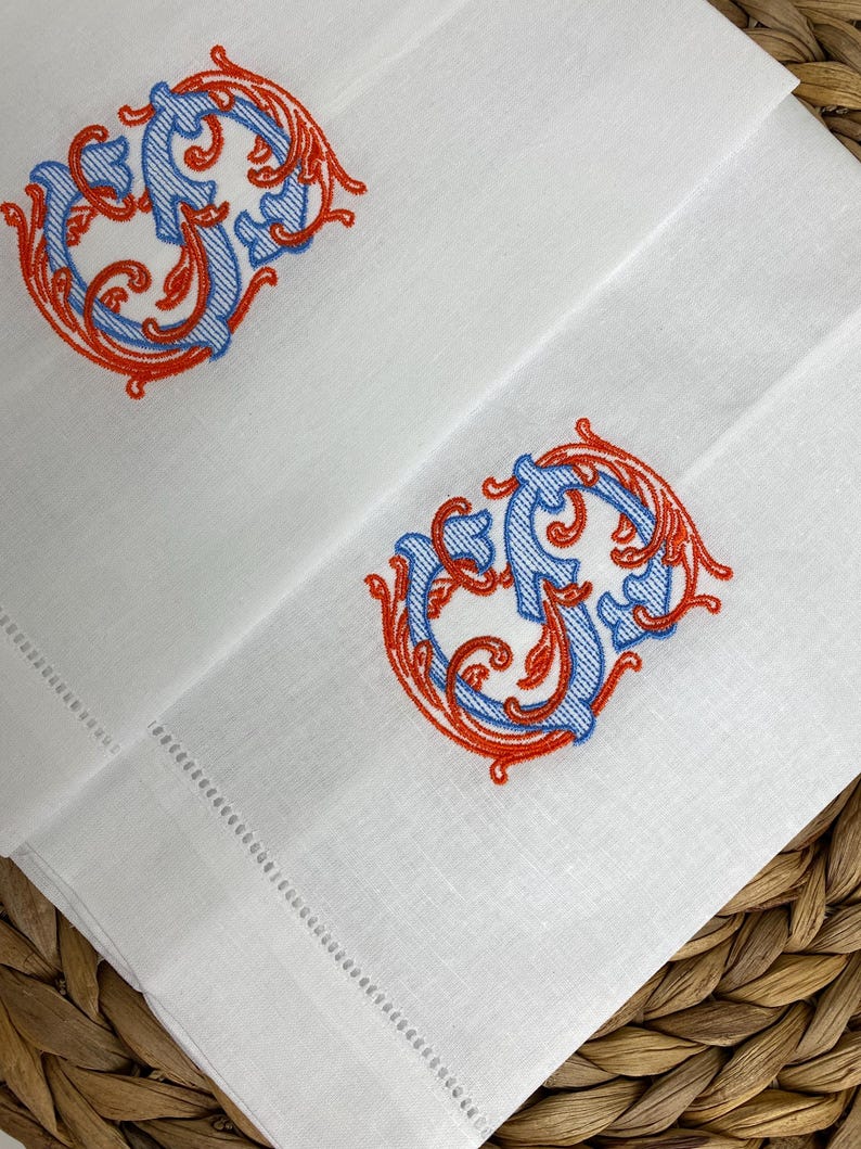 May include: Two white linen napkins featuring an embroidered monogram letter S in a decorative script style. The embroidery uses a combination of light blue thread for the letter and vibrant orange thread for the surrounding ornate scrollwork.
