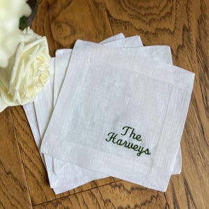 May include: A stack of white linen cocktail napkins with the embroidered text "The Harveys" in green. The napkins are square and have a subtle border. A white rose is visible in the background, resting on a wooden surface.