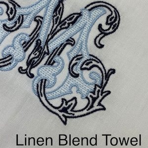 May include: A white linen blend towel featuring an embroidered monogram letter M in dark navy blue and light blue thread. The embroidery has a decorative, swirling scrollwork design. The text Linen Blend Towel is printed in black below.