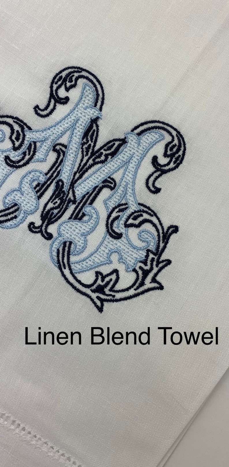 May include: A white linen blend towel featuring an embroidered monogram letter M in dark navy blue and light blue thread. The embroidery has a decorative, swirling scrollwork design. The text Linen Blend Towel is printed in black below.